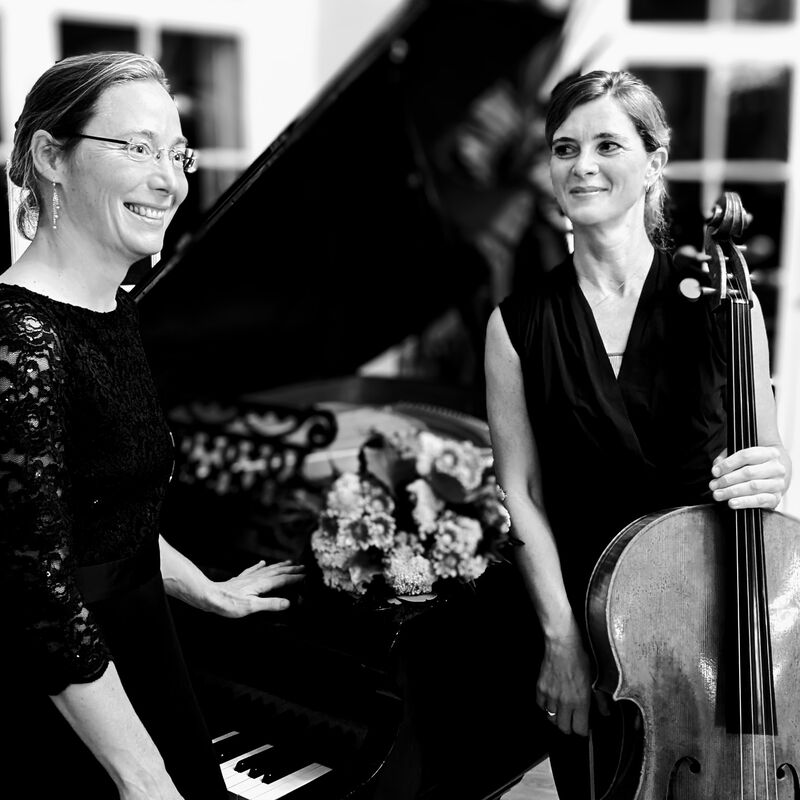 Das Schwarzweiß-Foto zeigt die Solocellistin der Hamburger Philharmonie Olivia Jeremias mit Pianistin Kerstin Mudra am aufgeklapten Flügel stehend